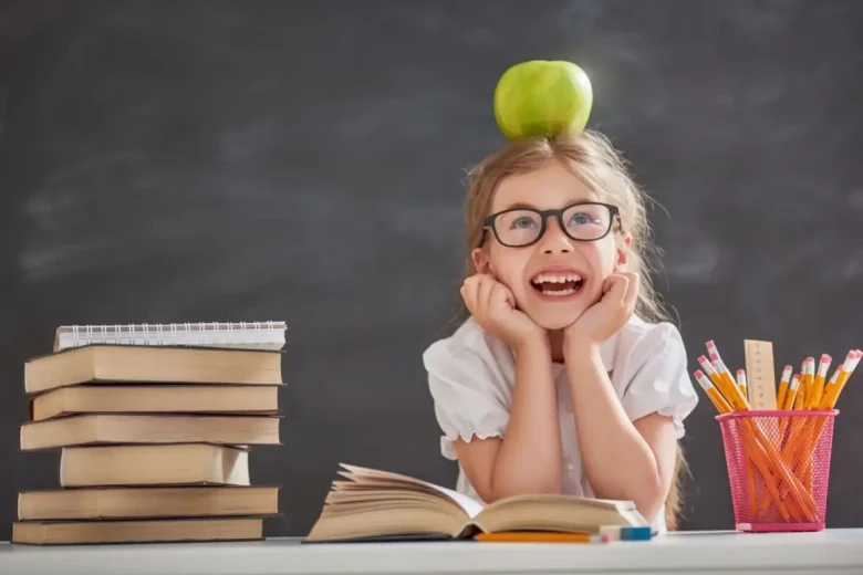 girl sat at a desk balancing an apple on her head and smiling, while reading a book