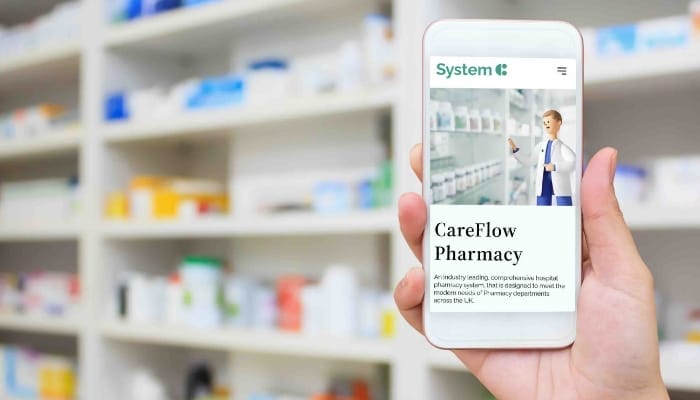 A picture of a pharmacist in a pharmacy setting, holding an electronic device displaying the System C CareFlow pharmacy solution.