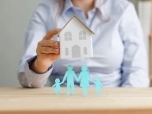 An image which shows a woman placing a house over silhouettes of a family.